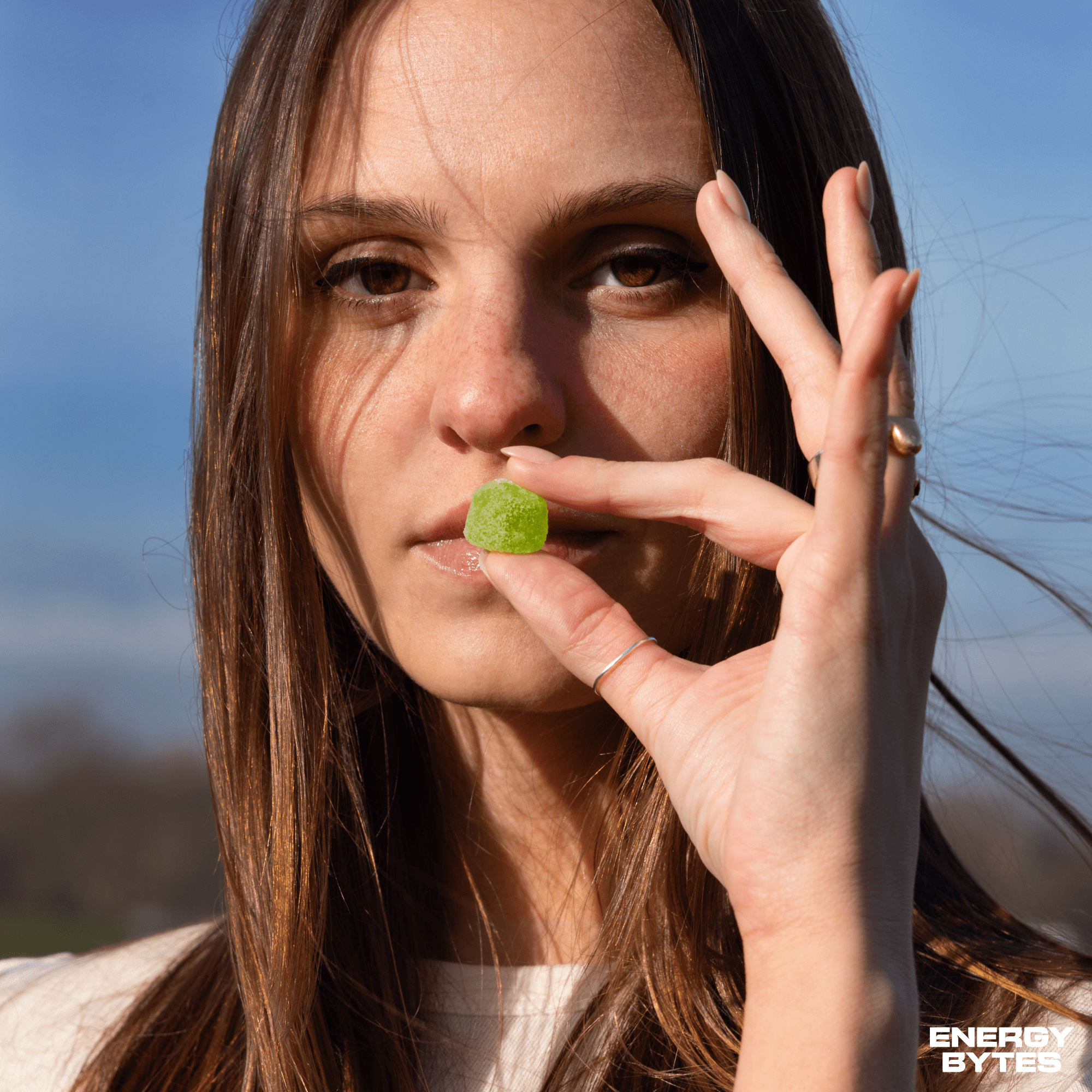 Woman holding up a single Atomic Apple Energy Bytes gummy before a clear sky backdrop, illustrating the zesty, energising snack option ideal for active lifestyles.