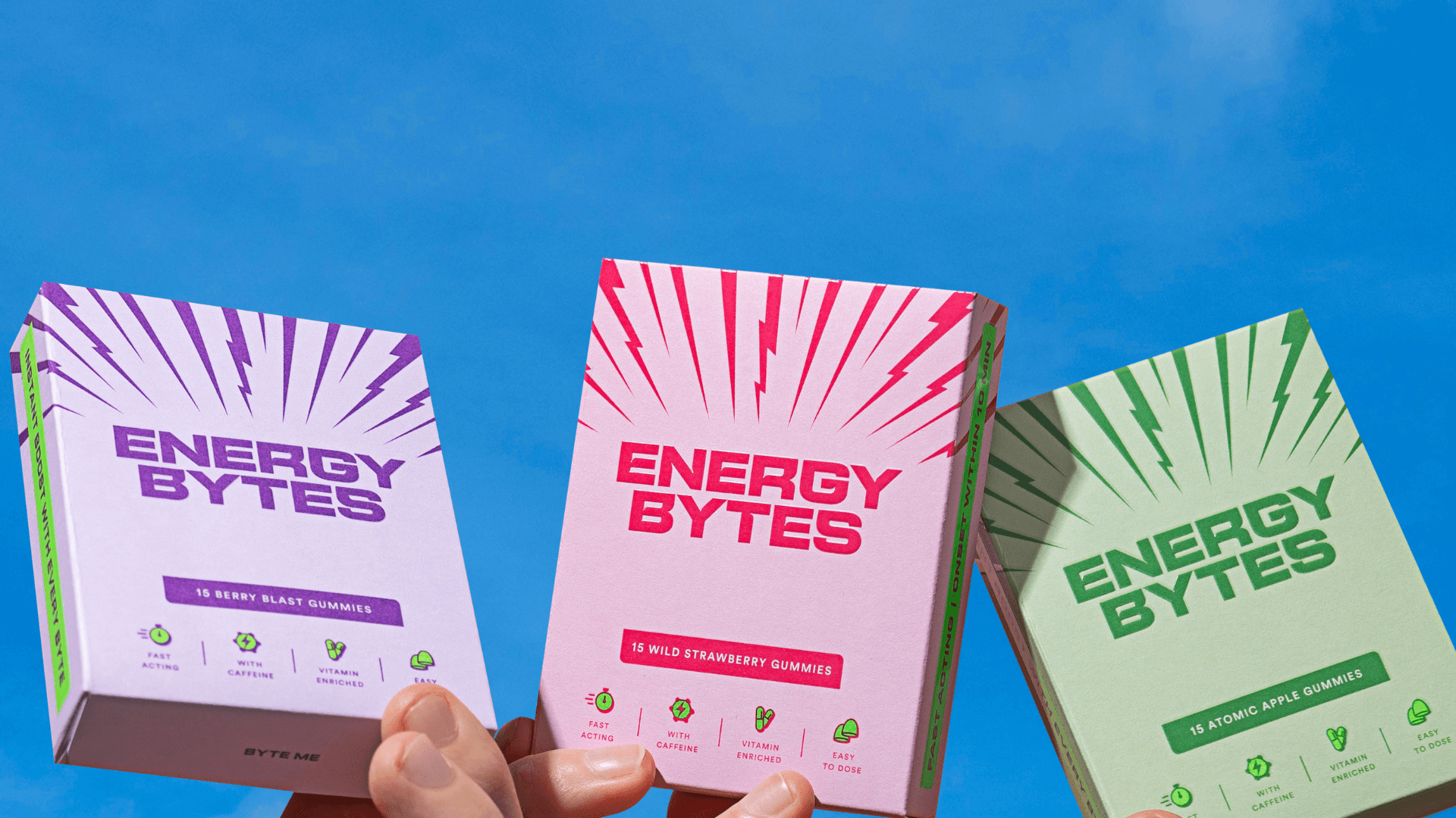 Hands holding up Energy Bytes gummy packets in Berry Blast, Wild Strawberry, and Atomic Apple flavours against a clear blue sky.