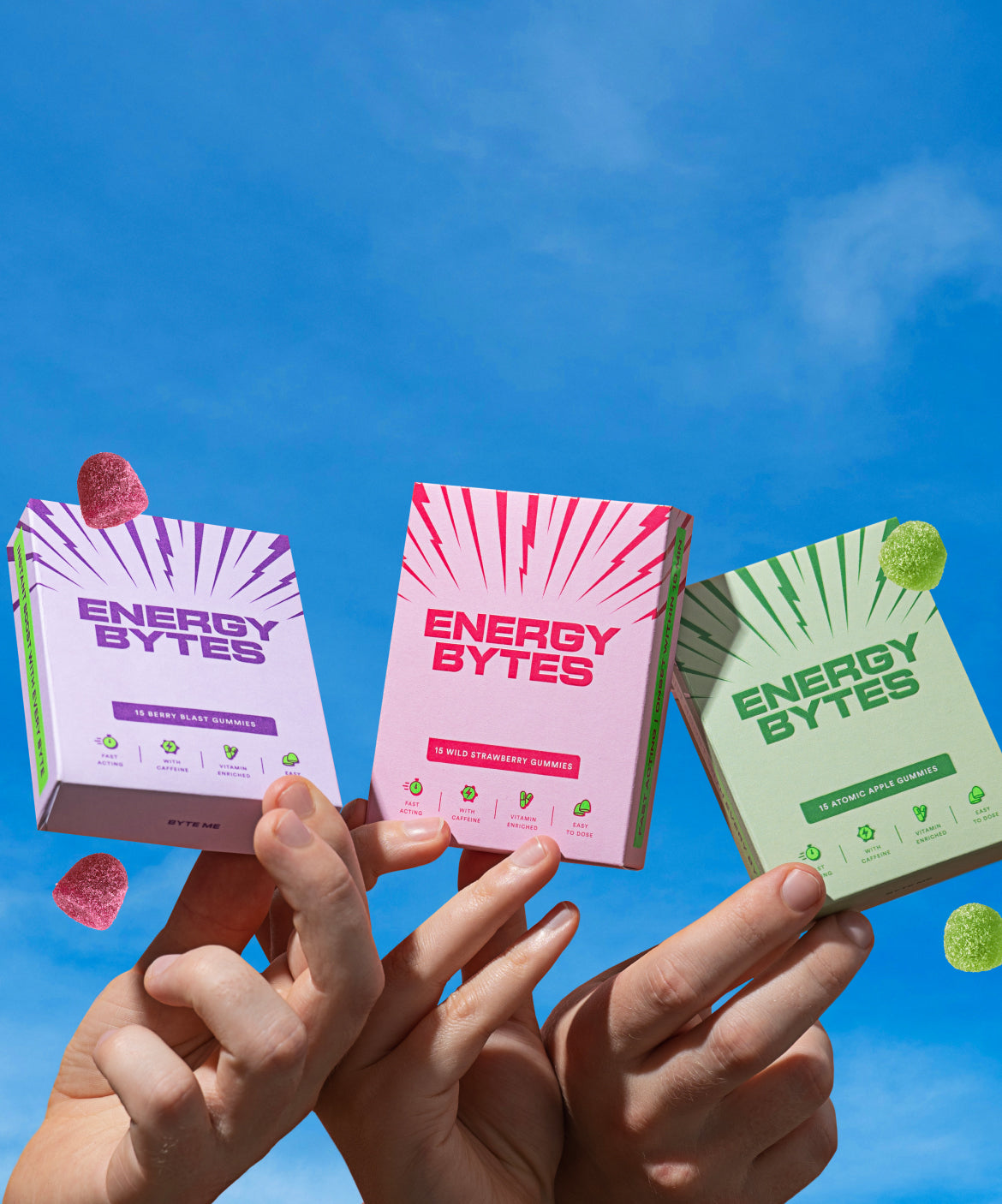 Hands holding up Energy Bytes gummy packets in Berry Blast, Wild Strawberry, and Atomic Apple flavours against a clear blue sky.